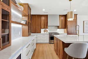 Kitchen with brown cabinetry, hanging light fixtures, light stone counters, light wood-type flooring, and stainless steel range