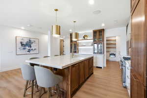 Kitchen featuring brown cabinets, a kitchen bar, glass insert cabinets, a kitchen island with sink, and light stone counters