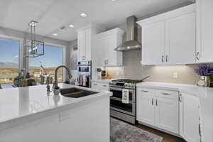 Kitchen featuring hanging light fixtures, white cabinets, wall chimney range hood, stainless steel appliances, and light stone counters