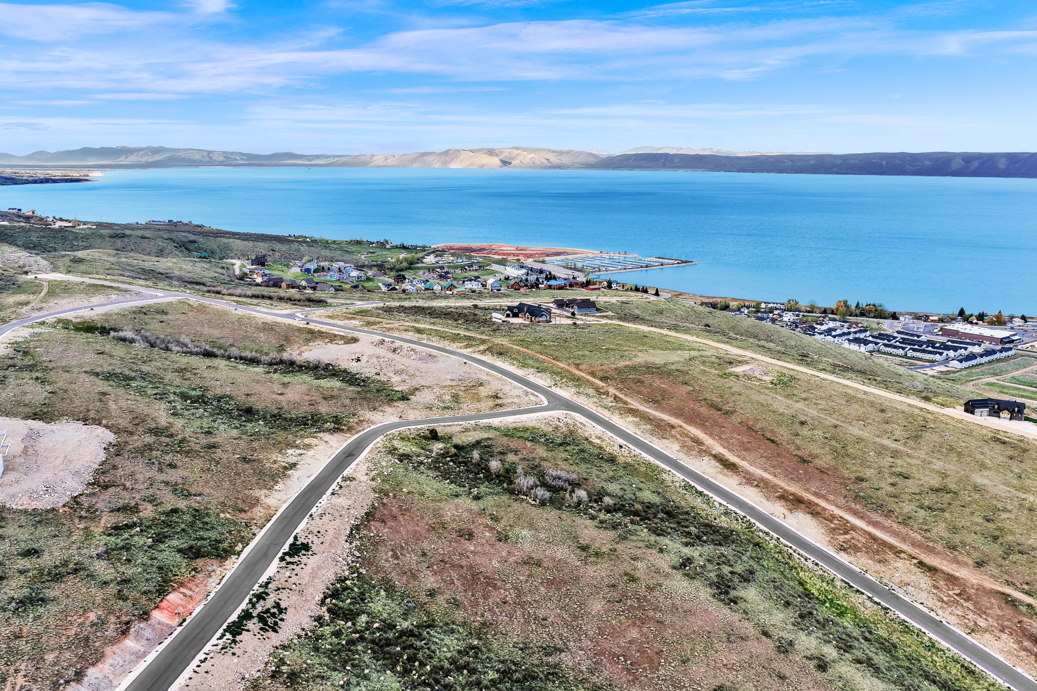 Expansive hillside land featuring paved roads, developing residential areas, and a prominent lake backdrop with distant mountains