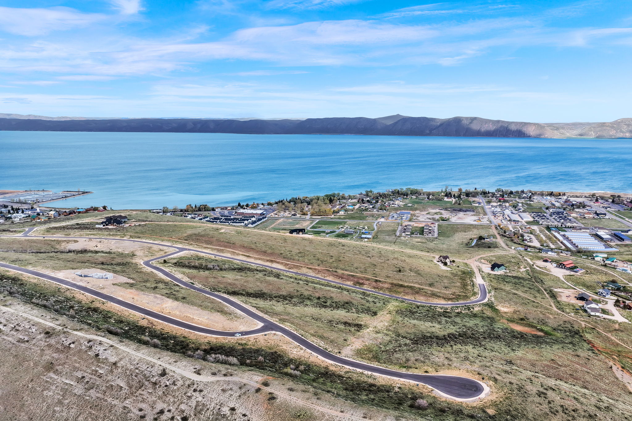 Expansive undeveloped land featuring paved roads and gentle slopes, offering proximity to a large body of water and distant mountainous terrain