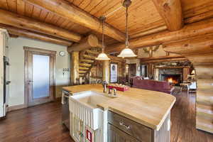 Kitchen with butcher block countertops, log walls, hanging light fixtures, a kitchen island with sink, and dark wood-style flooring