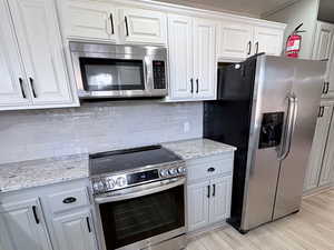 Kitchen featuring appliances with stainless steel finishes, light stone countertops, decorative backsplash, white cabinets, and light wood-style floors