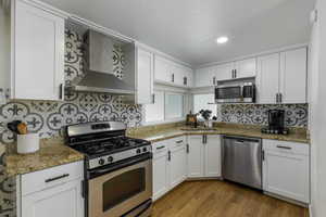 Kitchen with appliances with stainless steel finishes, wall chimney exhaust hood, white cabinets, dark wood-style flooring, and light stone countertops