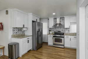 Kitchen with appliances with stainless steel finishes, wall chimney exhaust hood, light stone counters, light wood finished floors, and recessed lighting