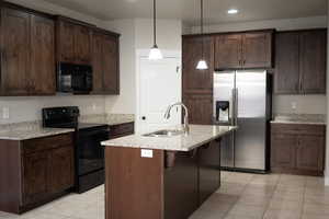 Kitchen featuring black appliances, pendant lighting, light stone countertops, dark brown cabinetry, and a center island with sink