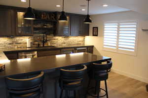 Bar area featuring light wood-type flooring, decorative light fixtures, glass insert cabinets, dishwasher, and dark brown cabinets