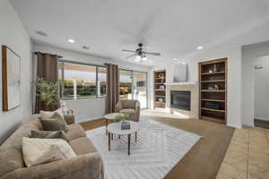 Living room with a tiled fireplace, built in shelves, ceiling fan, light tile patterned flooring, and light colored carpet