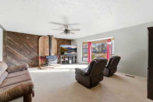Carpeted living room featuring a wood stove, wood walls, ceiling fan, and a textured ceiling