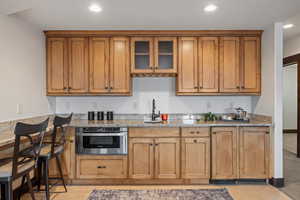 Kitchen with brown cabinetry, light stone counters, recessed lighting, glass insert cabinets, and stainless steel microwave
