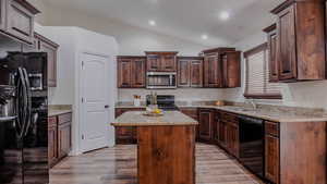 Kitchen with black appliances, a center island, vaulted ceiling, light stone counters, and dark brown cabinets