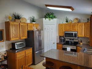 Kitchen with backsplash, appliances with stainless steel finishes, dark stone counters, and brown cabinetry