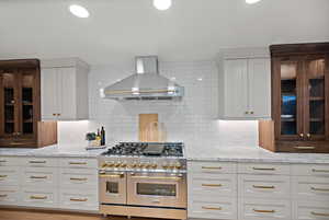 Kitchen with range with two ovens, wall chimney range hood, white cabinetry, light stone countertops, and recessed lighting