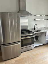 Kitchen with appliances with stainless steel finishes, wall chimney range hood, light wood finished floors, and light stone counters