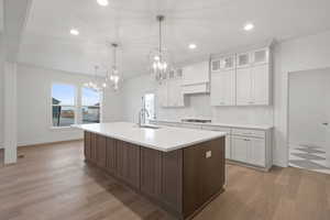 Kitchen featuring glass insert cabinets, light wood-type flooring, white cabinets, decorative light fixtures, and a center island with sink