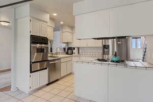 Kitchen featuring tile counters, appliances with stainless steel finishes, a warming drawer, a peninsula, and white cabinetry