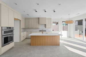 Kitchen with light stone countertops, stainless steel appliances, a kitchen island with sink, cream cabinets, and decorative light fixtures