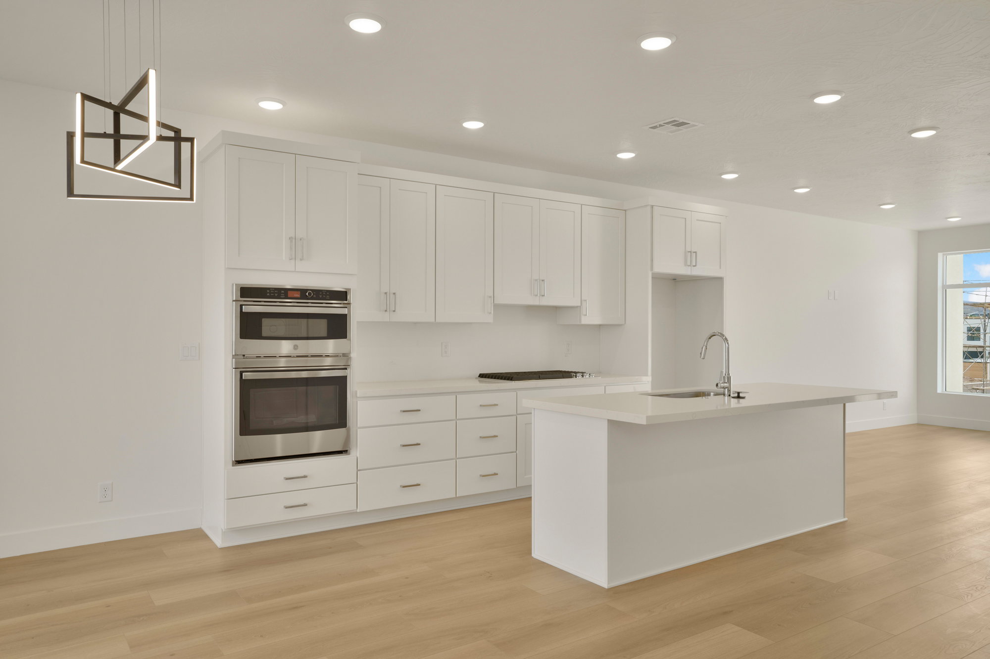Modern kitchen featuring white shaker-style cabinetry, a built-in stainless steel oven and microwave, light-toned wood-finish flooring, a contemporary island with an integrated sink, and recessed ceiling lighting
