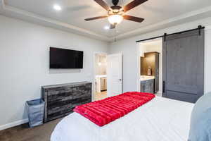 Bedroom featuring light colored carpet, a barn door, a ceiling fan, a tray ceiling, and ensuite bath
