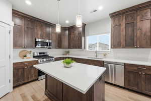 Kitchen featuring dark brown cabinetry, appliances with stainless steel finishes, decorative light fixtures, a kitchen island, and light stone countertops
