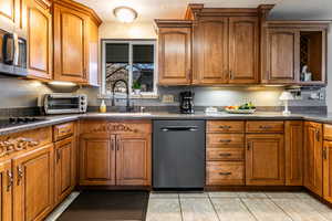 Kitchen with brown cabinetry, dishwasher, stainless steel microwave, light tile patterned floors, and black electric stovetop,  walk in pantry and hallway to front door
