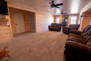 Living area with a wood stove, carpet floors, wooden walls, a textured ceiling, and a ceiling fan