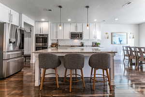 Kitchen featuring stainless steel appliances, a center island with sink, dark wood-style flooring, white cabinetry, and hanging light fixtures