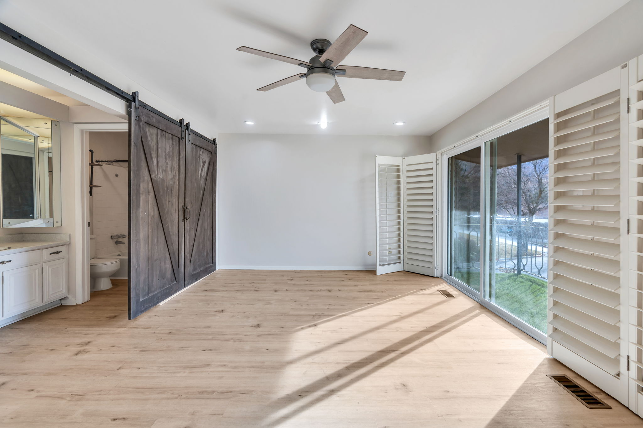 Bedroom featuring light wood-type flooring, access to outside, ceiling fan, a barn door, and ensuite bathroom
