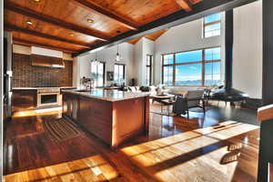 Kitchen with light stone countertops, hanging light fixtures, dark wood-style floors, wall chimney range hood, and high vaulted ceiling