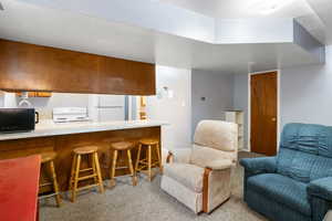 Mother-in-law basement apartment kitchen with light countertops, brown cabinetry, a breakfast bar, white appliances, and light-colored carpet.