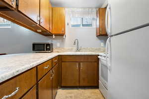 Mother-in-law basement apartment kitchen with light countertops, brown cabinetry, a breakfast bar, white appliances, and light-colored carpet.