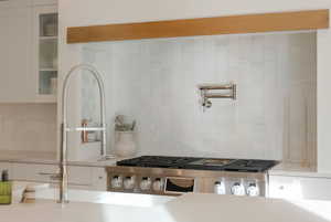 Kitchen view of pot filler, white cabinetry, ventilation hood, light stone countertops, and backsplash