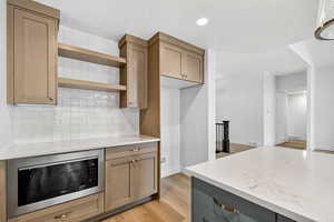 Kitchen with stainless steel microwave, open shelves, light stone countertops, light wood-type flooring, and decorative backsplash