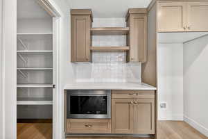Kitchen featuring stainless steel microwave, light stone countertops, light wood finished floors, open shelves, and decorative backsplash