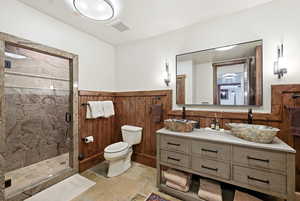 Full bathroom featuring double vanity, a shower stall, stone tile floors, and a wainscoted wall