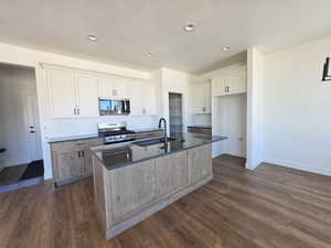 Two tone kitchen with stainless steel appliances, a center island with sink, dark wood-style flooring, two tone cabinets, and dark stone countertops