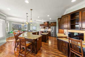 Kitchen with light stone countertops, ornamental molding, pendant lighting, dark brown cabinetry, and dark wood-style flooring