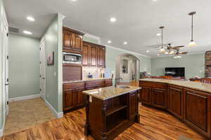 Kitchen with arched walkways, ceiling fan, hanging light fixtures, a kitchen island with sink, and light stone countertops