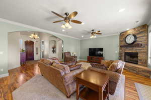 Living room featuring wood finished floors, a stone fireplace, ceiling fan, ornamental molding, and arched walkways