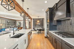Kitchen showcasing functional excellence and custom design, featuring elegant pendant lighting, light wood-style flooring, an open-concept layout, a bespoke range hood, and luminous light-stone countertops.