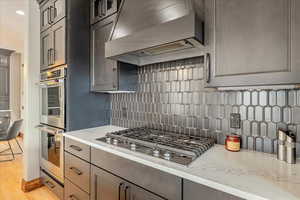 Kitchen featuring a premium range hood above a professional gas range, stainless steel appliances, a tasteful designer backsplash, light stone countertops, and refined cabinetry.