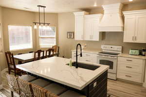 Kitchen with white range with electric stovetop, light stone counters, custom range hood, white cabinets, and a textured ceiling