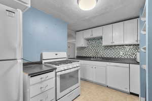 Kitchen with white appliances, white cabinets, dark countertops, backsplash, and a textured ceiling