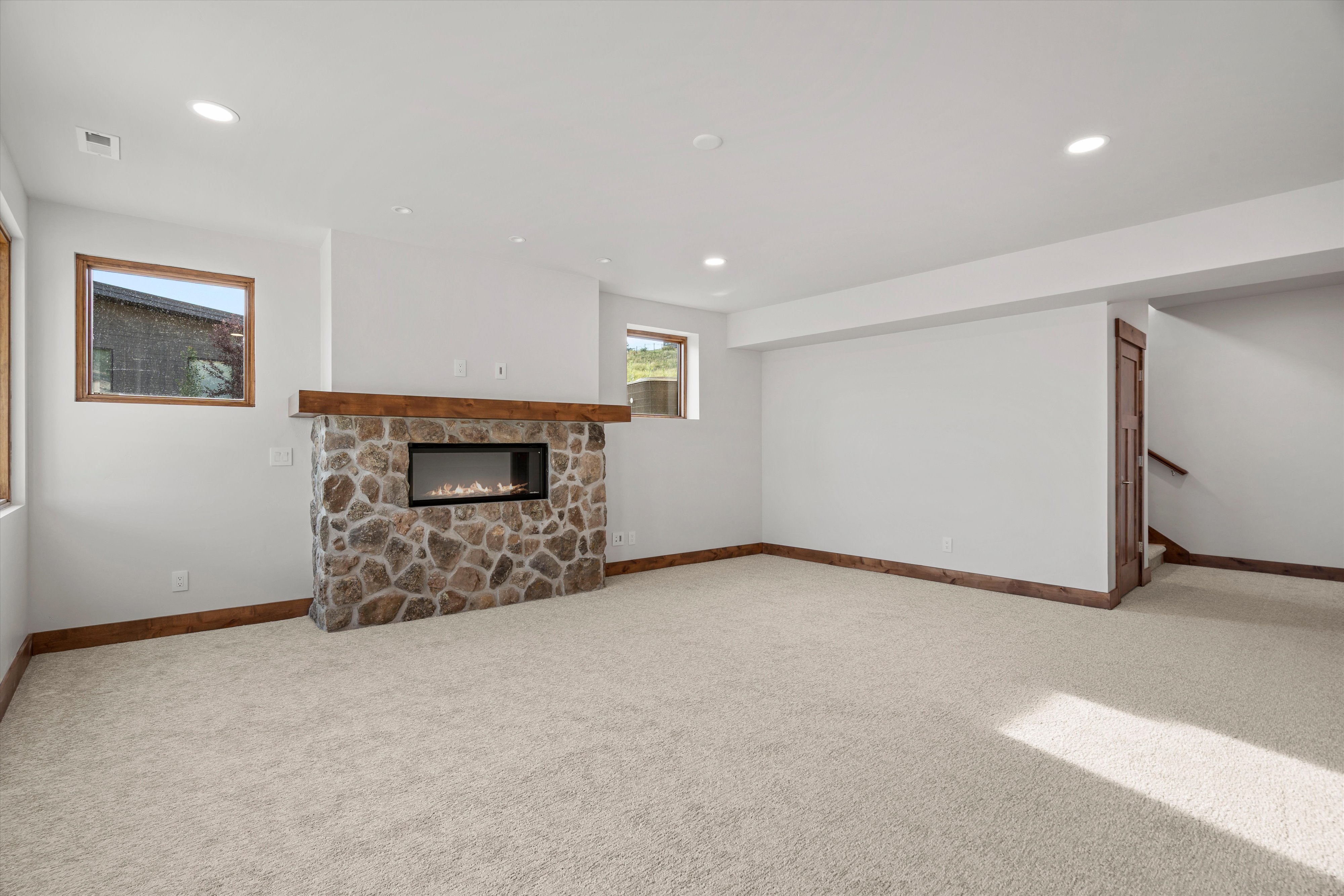 Spacious room featuring a stone-clad fireplace with a natural wood mantel, recessed lighting, neutral carpet flooring, and wood-trim windows