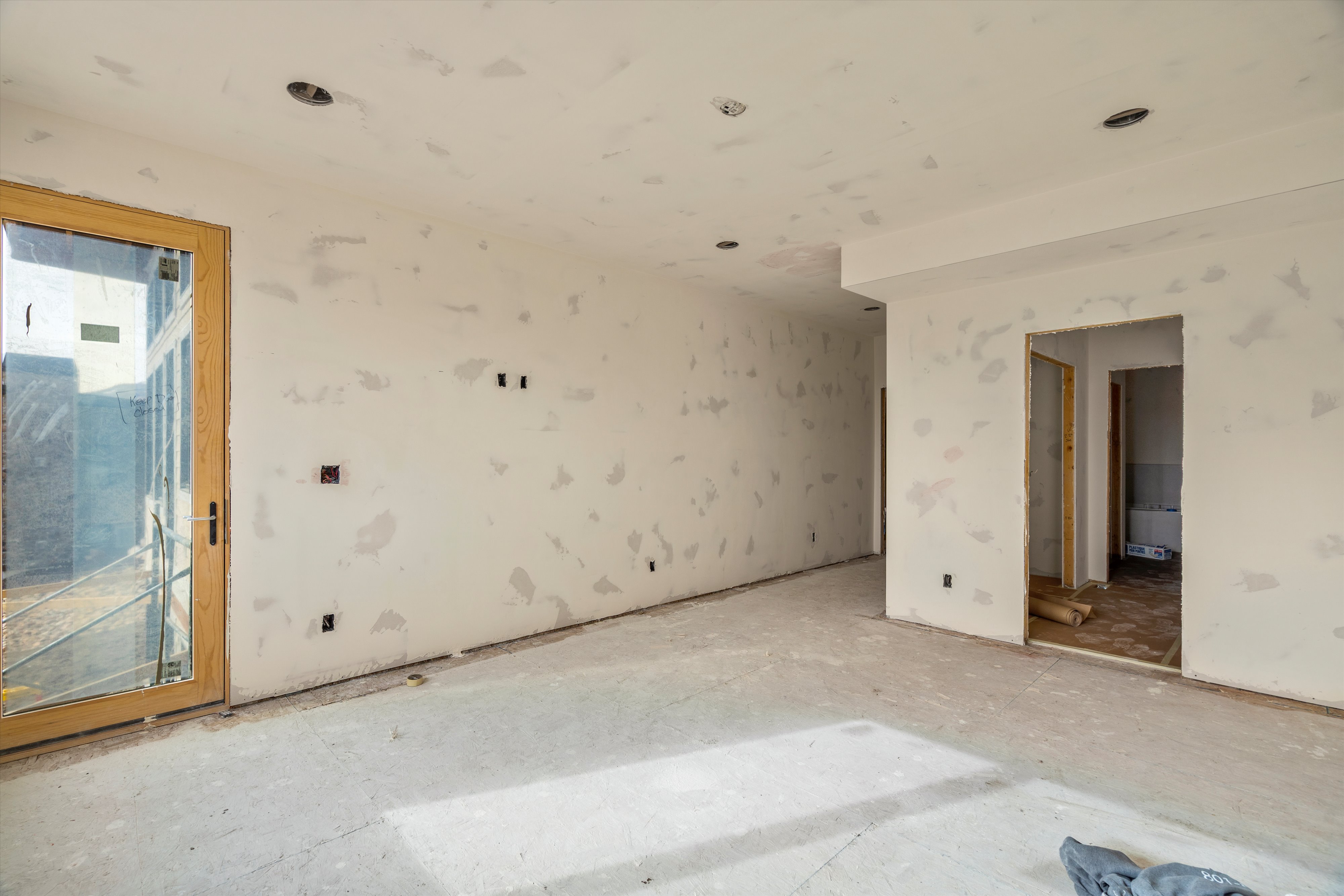 Interior space showcasing a large glass door with a light wood frame, multiple recessed light fixtures, and raw drywall finishes