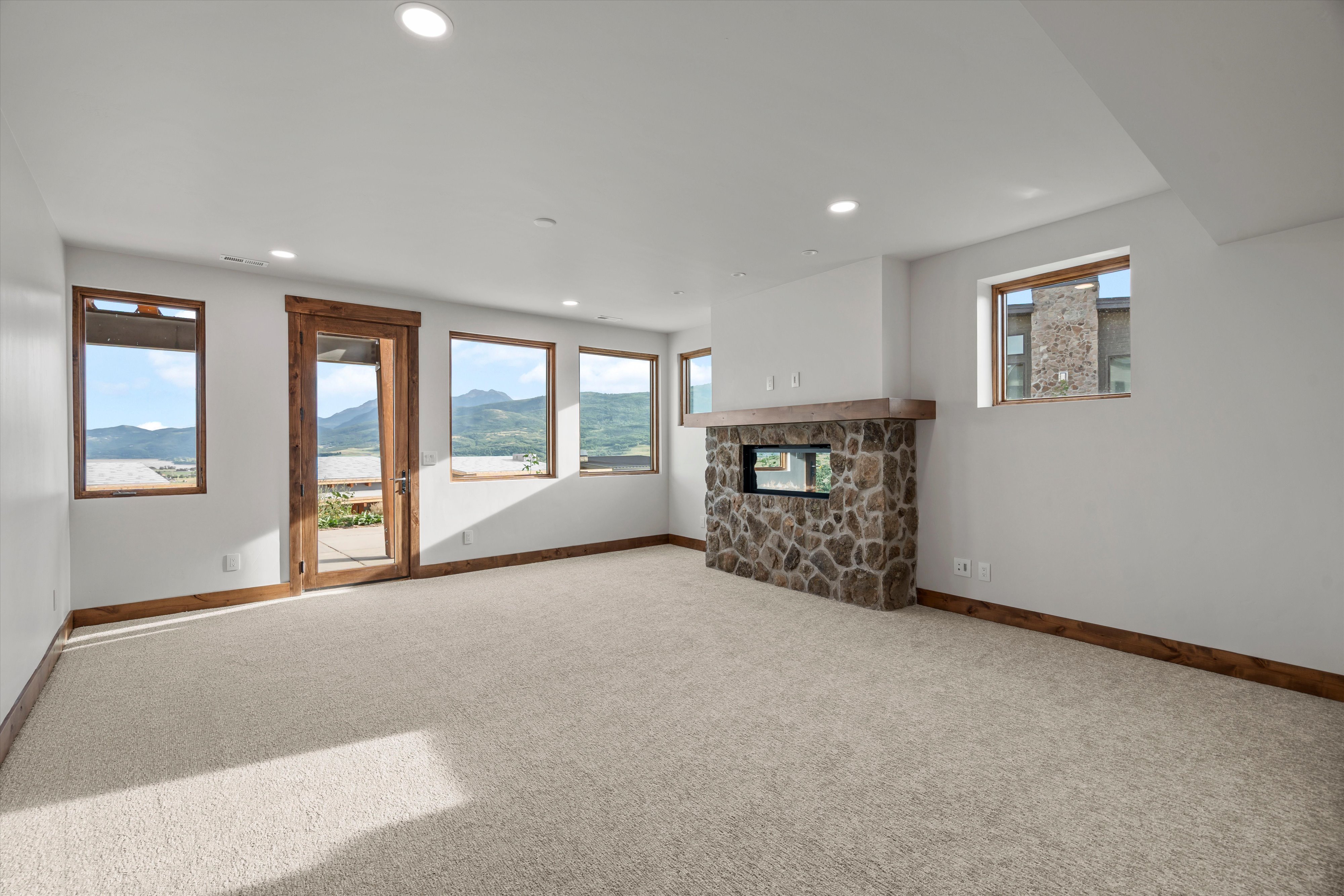 Spacious room featuring light-colored carpeting, a stone-clad fireplace with a wood mantle, and multiple windows with wood trim