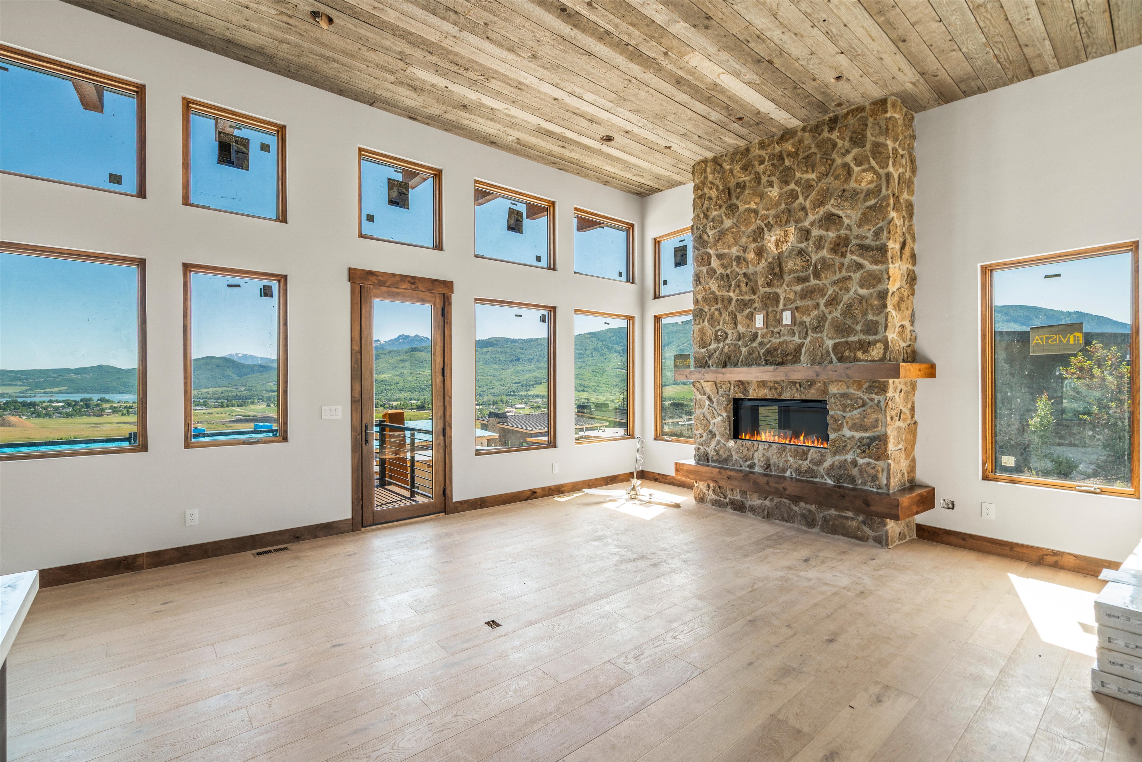 Spacious room featuring a floor-to-ceiling stone fireplace with a wood mantle and electric insert, multiple large windows with wood trim, and a wood-finish ceiling