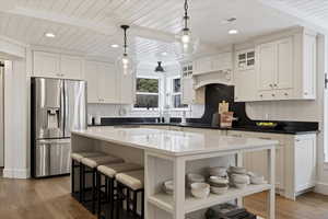 Kitchen with glass insert cabinets, stainless steel fridge with ice dispenser, dark stone countertops, light wood-style flooring, and decorative light fixtures