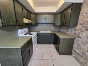 Unit #2- Kitchen featuring brick wall, white electric stove, light countertops, under cabinet range hood, and light tile patterned floors