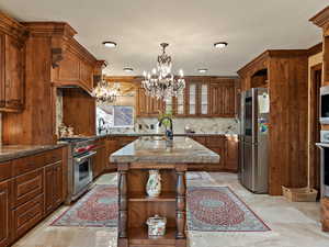 Kitchen featuring open shelves, an island with sink, stainless steel appliances, decorative light fixtures, and brown cabinetry